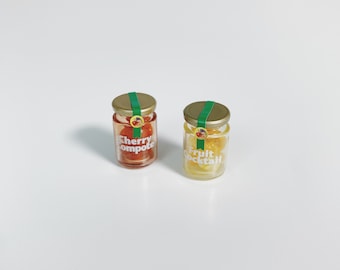 Può includere: Due barattoli di vetro in miniatura con coperchi in metallo dorato e sigilli di carta verde. Uno contiene composta di ciliegie rossa con testo bianco, l'altro macedonia di frutta gialla con testo bianco. Entrambi hanno un piccolo logo.