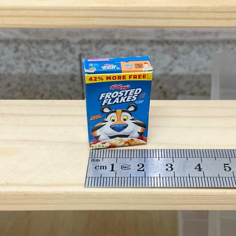 May include: Miniature Kellogg's Frosted Flakes cereal box. The box is blue with the Frosted Flakes logo and Tony the Tiger. The box reads "42% MORE FREE". A ruler is next to the box, showing measurements in centimeters.