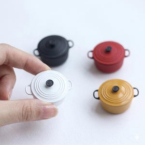 May include: Miniature colorful cooking pots in white, black, red, and yellow. Each pot has a matching lid with a black knob and small side handles. The white pot is held by a hand. These are likely for a dollhouse or miniature collection.