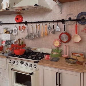 May include: A white kitchen with a black metal rack above the stove. The rack holds various kitchen utensils, including a red pan, a white ladle, and a yellow wooden spoon. The stove has four burners and a white oven. There are white cabinets on either side of the stove. The countertop is made of wood and has a variety of kitchen items on it, including a red bowl, a green plant, and a wooden box labeled "Natural Herb".