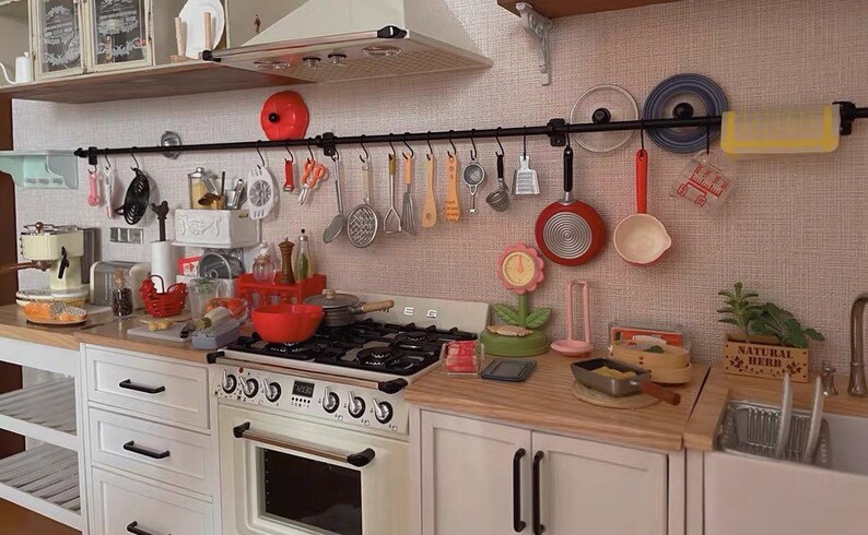 May include: A white kitchen with a black metal rack above the stove. The rack holds various kitchen utensils, including a red pan, a white ladle, and a yellow wooden spoon. The stove has four burners and a white oven. There are white cabinets on either side of the stove. The countertop is made of wood and has a variety of kitchen items on it, including a red bowl, a green plant, and a wooden box labeled "Natural Herb".