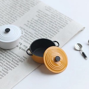 May include: Miniature white and yellow cooking pots with black handles and lids, along with two small silver spoons. The pots are on a newspaper background, suggesting a culinary or kitchen theme.