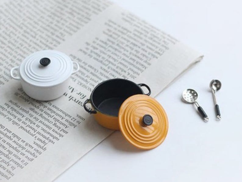 May include: Miniature white and yellow cooking pots with black handles and lids, along with two small silver spoons. The pots are on a newspaper background, suggesting a culinary or kitchen theme.