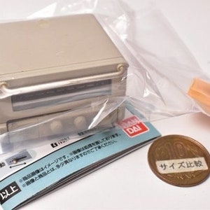 May include: A miniature beige toaster oven toy encased in a clear plastic bag, featuring a front handle, two control knobs, and a glass window. A small orange accessory piece, a printed instruction manual, and a Japanese ten-yen coin are included.