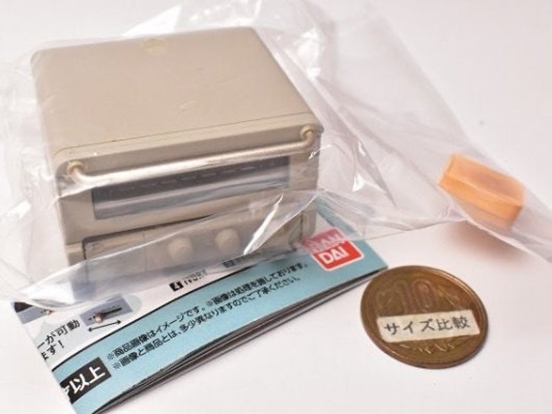 May include: A miniature beige toaster oven toy encased in a clear plastic bag, featuring a front handle, two control knobs, and a glass window. A small orange accessory piece, a printed instruction manual, and a Japanese ten-yen coin are included.