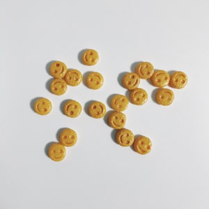May include: A collection of small, round, yellow smiley face decorations scattered on a white surface. Each decoration features a smiling face with two eyes and a curved mouth. The decorations are evenly spaced and appear to be made of a solid material.