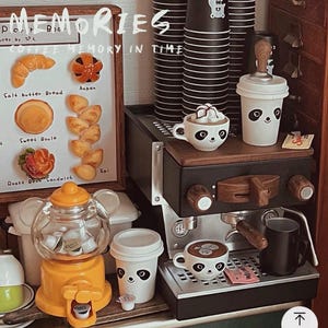Pode incluir: Uma cena de caf&eacute; em miniatura com uma m&aacute;quina de caf&eacute; expresso preta, x&iacute;caras com rostos de panda e uma m&aacute;quina de chicletes. Uma placa exibe miniaturas de produtos de panifica&ccedil;&atilde;o. O texto "MEMORIES" e "COFFEE MEMORY IN TIME" s&atilde;o vis&iacute;veis.