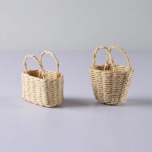 Puede incluir: Dos cestas tejidas con asas. La cesta más pequeña es rectangular y la más grande es redonda. Ambas cestas están hechas de paja natural.