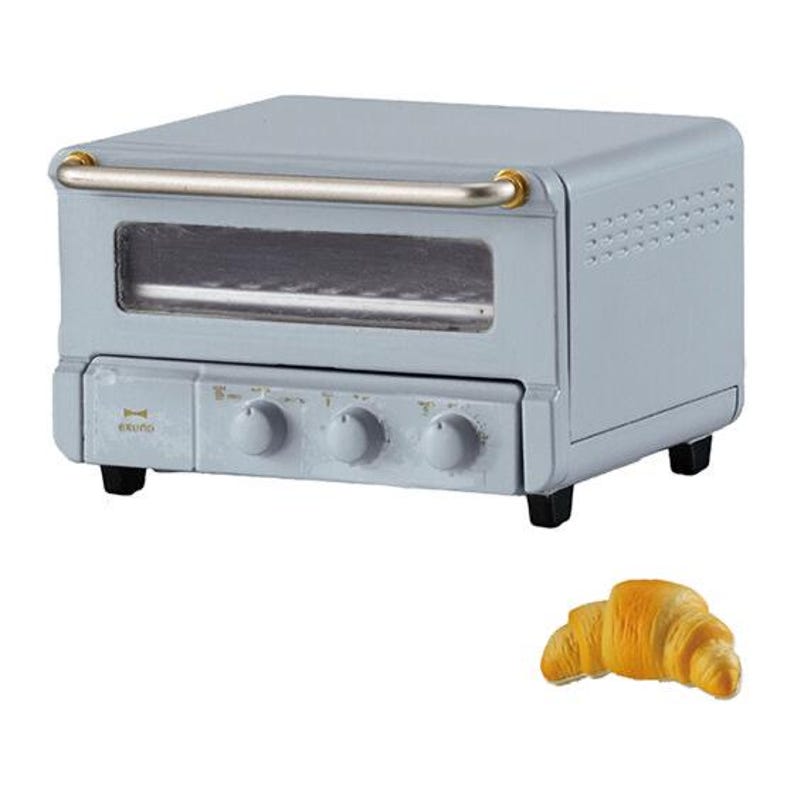 May include: A light blue toaster oven with a horizontal silver handle, a glass viewing window, and three control knobs on the front panel. The brand name Bruno is printed in gold on the left side. A single golden-brown croissant is placed below.