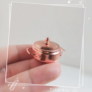 May include: A miniature copper pot with a lid and two handles. The pot is small enough to fit in the palm of a hand.