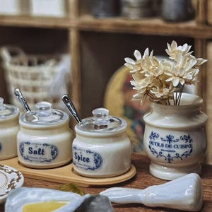 Può includere: Set da cucina in miniatura con barattoli di spezie color crema con etichette "Salt" e "Spice" con cucchiaini d'argento, un piccolo vaso con fiori bianchi e un cucchiaio da portata. I barattoli e il vaso hanno decorazioni floreali blu.
