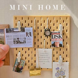 Può includere: Un pannello forato beige con vari oggetti in miniatura attaccati, tra cui una piccola borsa dorata, una rivista, un giornale, un orologio, una cartolina e una rivista. Il pannello forato ha le parole "MINI HOME" stampate in alto.