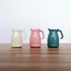 May include: Three ceramic teapots in cream, pink, and teal. Each teapot has a handle and a small spout. The teapots are displayed on a wooden surface against a white background. The teapots are a modern design.