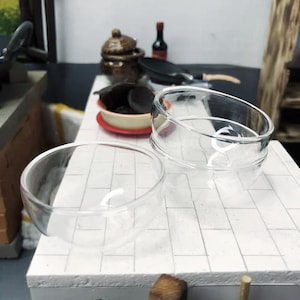 Puede incluir: Dos cuencos de vidrio transparente sobre una superficie de azulejos blancos. Los cuencos son redondos y tienen una ligera curva. Ambos están vacíos.