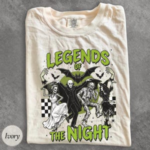 May include: An ivory t-shirt featuring a graphic print with the text "LEGENDS OF THE NIGHT" in green. The design includes a skeleton in a dress, a vampire, a zombie, and a witch, all positioned beneath a large bat against a green circle background.