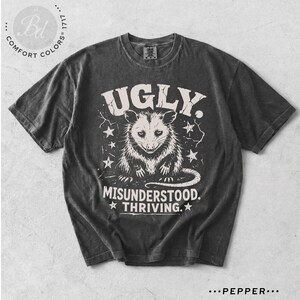 May include: A charcoal gray short-sleeve t-shirt featuring a graphic of an opossum centered on the chest. The text reads UGLY. MISUNDERSTOOD. THRIVING. The design includes small star accents and a distressed, weathered appearance on the fabric.