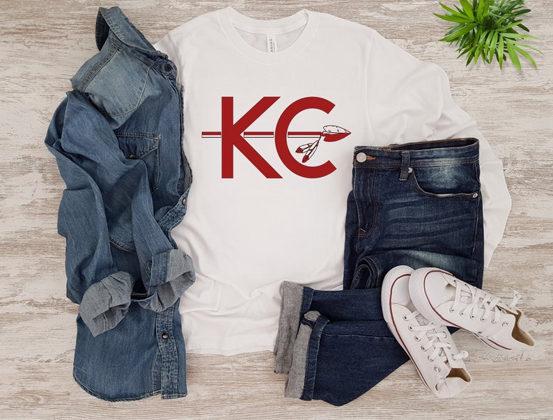 May include: White long-sleeve shirt with a red "KC" logo and a red arrow with feathers. The shirt is laid out on a wooden surface with a pair of blue jeans and white sneakers.