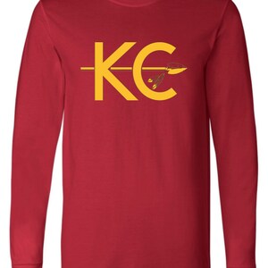 May include: Red long-sleeve t-shirt with a yellow graphic of the letters "KC" with an arrow through the "C".