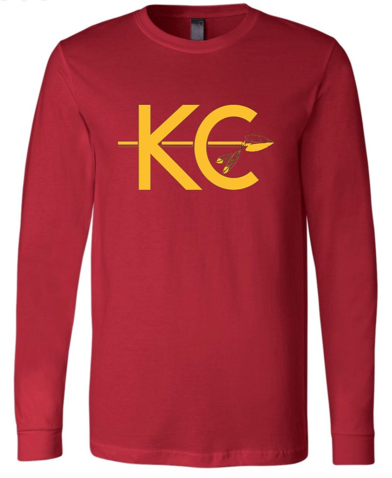 May include: Red long-sleeve t-shirt with a yellow graphic of the letters "KC" with an arrow through the "C".