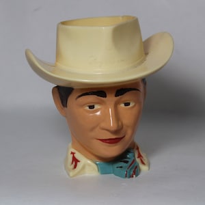 May include: A white plastic cowboy hat with a smiling face and a blue and red neckerchief. The hat has the word "Roy" written on it.