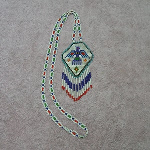 Puede incluir: Un collar de cuentas con un colgante de pájaro blanco, azul, rojo y verde. El colgante es de forma geométrica con una franja de cuentas.