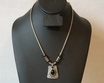 May include: A silver chain necklace with a black stone pendant. The pendant is a rectangular shape with a hammered texture and a black oval stone in the center. The chain is a snake chain style.