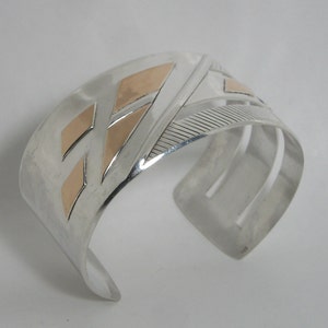 May include: Wide silver cuff bracelet with geometric gold-colored accents. The bracelet features a series of cut-out lines and diamond shapes. The cuff is a bold statement piece.