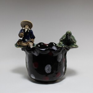 May include: A black ceramic planter with white and red polka dots. The planter has a ruffled edge and features a small figurine of a man sitting on a turtle.