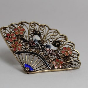 May include: A silver filigree brooch in the shape of a fan with two birds and pink flowers. The birds are black and white with blue and red accents. The fan is decorated with a blue enamel eye.