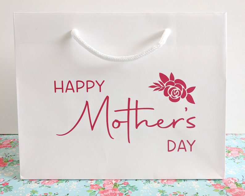 White gift bag with a rope handle. The bag has pink text that says "Happy Mother's Day" with a stylised rose and leaves.