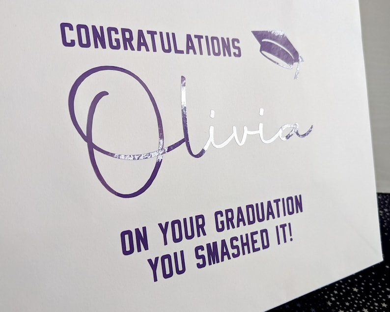 A white gift bag with purple text that reads "Congratulations Olivia on your graduation you smashed it!" with a purple graduation cap icon.