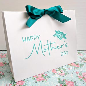 White gift bag with teal text and ribbon. The bag says Happy Mother's Day with a floral motif above.