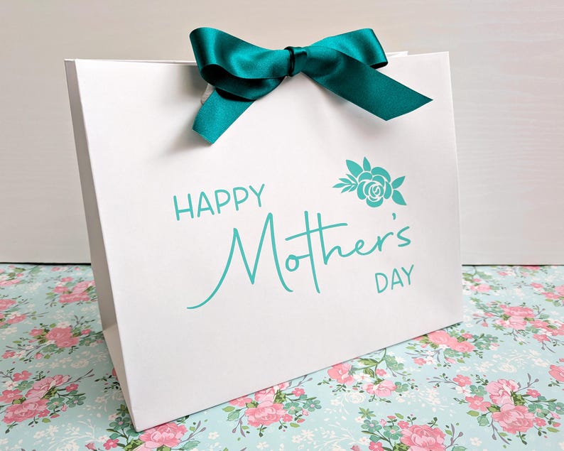 White gift bag with teal text and ribbon. The bag says Happy Mother's Day with a floral motif above.