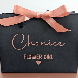 Black gift bag with a rose gold ribbon and bow. The bag has the name "Chonice" and the words "Flower Girl" in rose gold.
