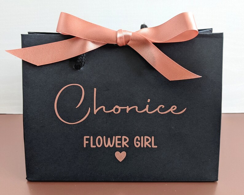 Black gift bag with a rose gold ribbon and bow. The bag has the name "Chonice" and the words "Flower Girl" in rose gold.