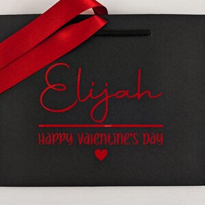 A black gift bag with red ribbon and text. The name is in large text with a Valentine's day message below.