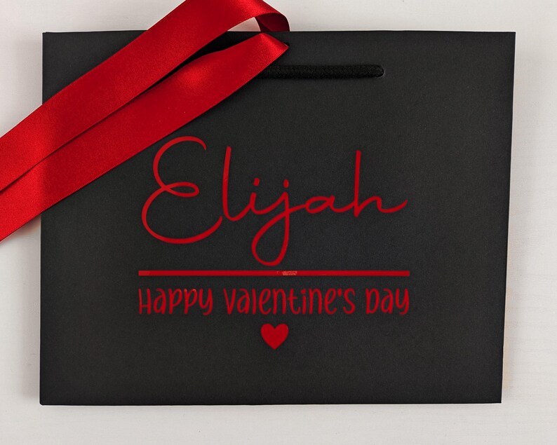 A black gift bag with red ribbon and text. The name is in large text with a Valentine's day message below.
