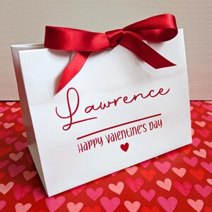 A white gift bag with red ribbon. It is personalised in red vinyl with a name and Happy Valentine's Day below in smaller text.