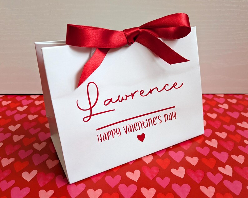 A white gift bag with red ribbon. It is personalised in red vinyl with a name and Happy Valentine's Day below in smaller text.