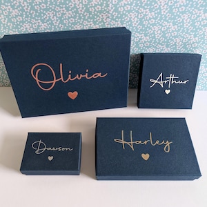 May include: Four rectangular navy blue gift boxes of varying sizes, each featuring a name in metallic script lettering and a small heart icon. The names Olivia, Arthur, Dawson, and Harley are printed in rose gold or gold foil on the lids.