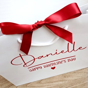 White gift bag with a red satin ribbon bow. The bag has the name "Danielle" and the text "Happy Valentine's Day" in red.