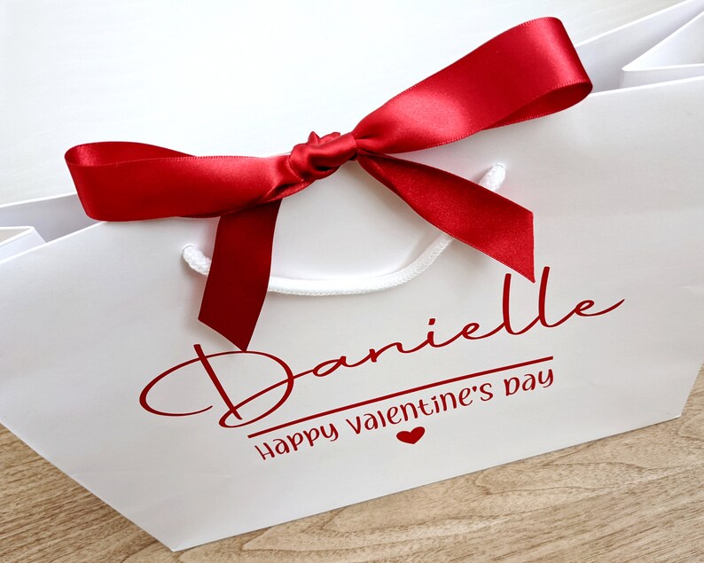 White gift bag with a red satin ribbon bow. The bag has the name "Danielle" and the text "Happy Valentine's Day" in red.