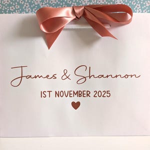 White gift bag with the bride and groom's names and their wedding date below. The text and ribbon are rose gold.