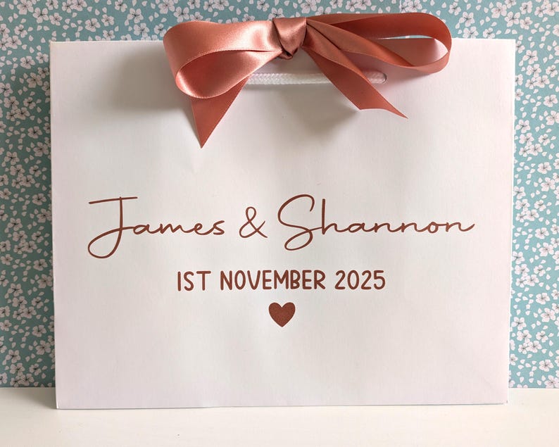 White gift bag with the bride and groom's names and their wedding date below. The text and ribbon are rose gold.