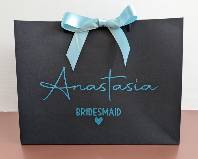 Black gift bag with a light blue bow and the text "Anastasia Bridesmaid" in teal blue lettering with a small teal heart.
