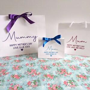 Three white gift bags with ribbon bows. The bags have personalised messages for "Mummy", "Mum", and "Mam" with the text "Happy Mother's Day" and a personalised message.