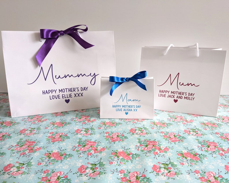 Three white gift bags with ribbon bows. The bags have personalised messages for "Mummy", "Mum", and "Mam" with the text "Happy Mother's Day" and a personalised message.