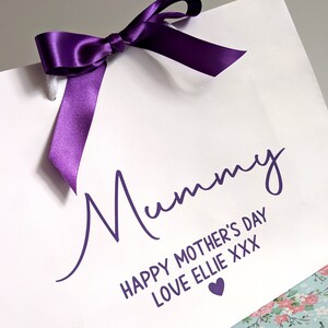 White gift bag with a purple ribbon and the text "Mummy Happy Mother's Day Love Ellie XXX" in purple lettering with a small purple heart.