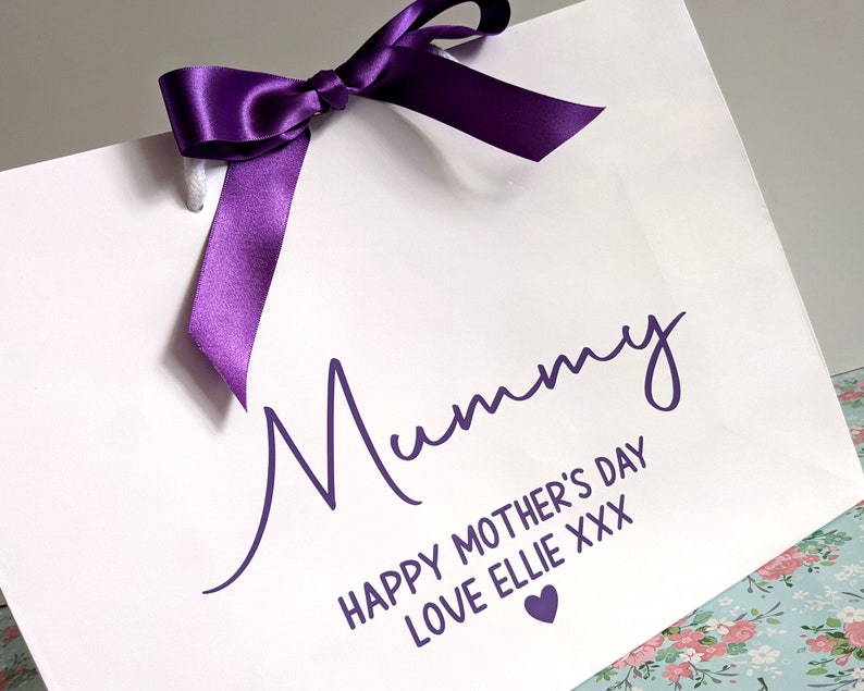 White gift bag with a purple ribbon and the text "Mummy Happy Mother's Day Love Ellie XXX" in purple lettering with a small purple heart.