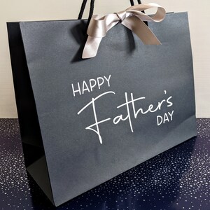 A black gift bag with a white satin ribbon and the text "Happy Father's Day" printed on the front.